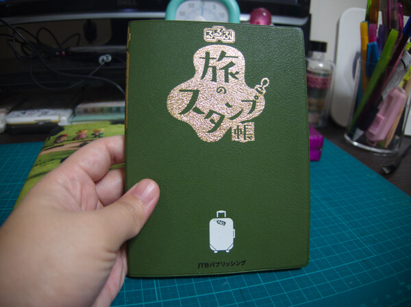 Small green book that reads 'Travel Stamp Book' in gold and a small white rolling suitcase at the bottom