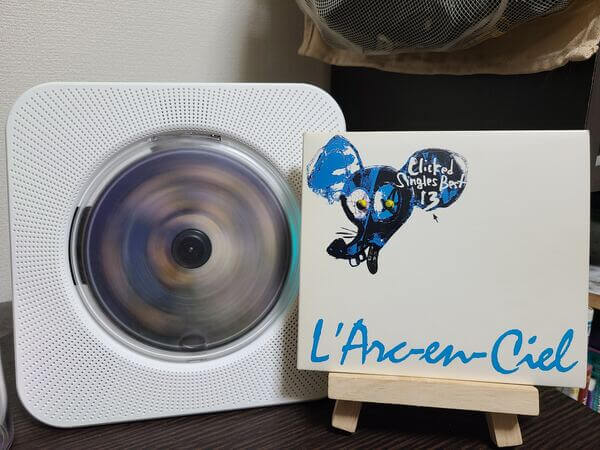 Blue and tan disc spinning in white CD player. Beside it is the album case for L'arc en Ciel's Clicked Best Singles 13