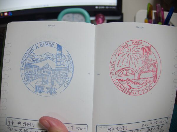 Small book held open with a blue stamp on the left of Ken-O Expressway's Atsugi Parking Area with Iiyama Onsen building, and a red stamp on the right of fireworks in the sky over a suspension bridge. A salmon is leaping in the water and the text says it is for the Ayu (Sweetfish) Festival