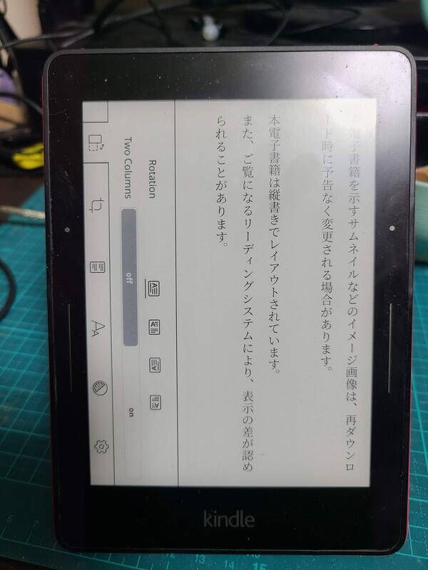 A Kindle Voyage using KOReader with the Japanese book loaded onto it as an EPUB. The text is displayed vertically from right-to-left, and on the left side of the screen is the UI rotated 90 degrees