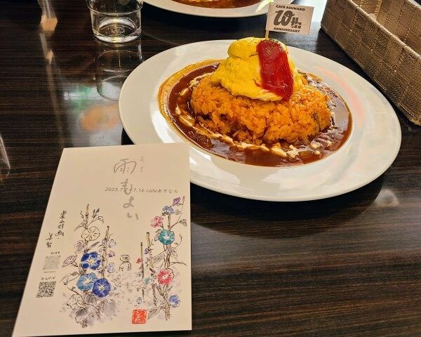 Rounded dome of omelettes surrounded by tomato sauce with a flag sticking out of the top. A postcard in the foreground leans against the plate with artwork of morning glories.