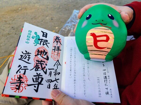 Person holding up a green daruma painted to look like a snake next to a goshuin (large kanji stamp) for Yugyoji