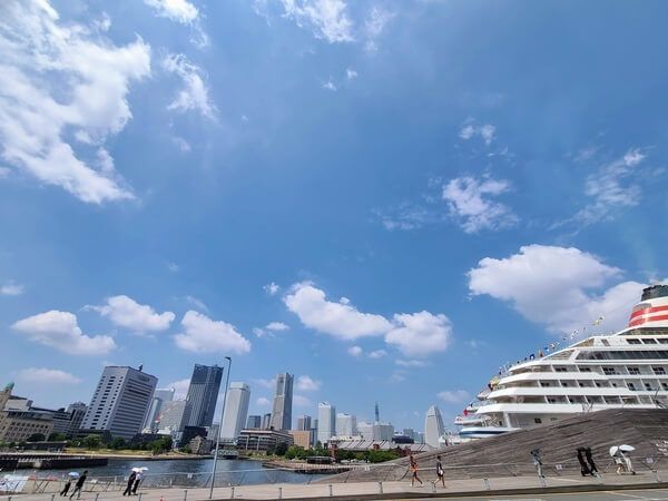 Yokohama's Minato Mirai skyline from across the harbor, with signatures like Landmark Tower, the Cosmo World wheel, and the Akarenga Warehouse. In the foreground is the Asuka II, a huge luxury liner, waiting at the Osanbashi pier