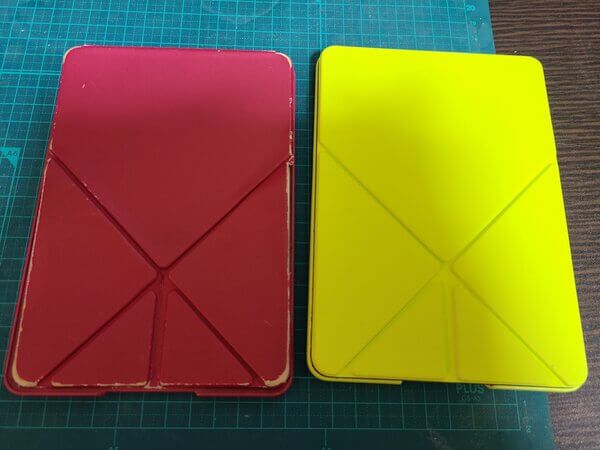 Two cases for the Kindle Voyage laid side-by-side on a desk. On the left is a battered and cracked pink case, and on the right is a yellow case holding the Voyage itself