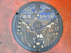 Colorful metal manhole cover for Shonan Hiratsuka. It depicts the Hiratsuka tanabata festival with the huge decorations put up around town and crowds of people attending the festival. In the background is the Sagami Bay with a yacht and sea birds flying around